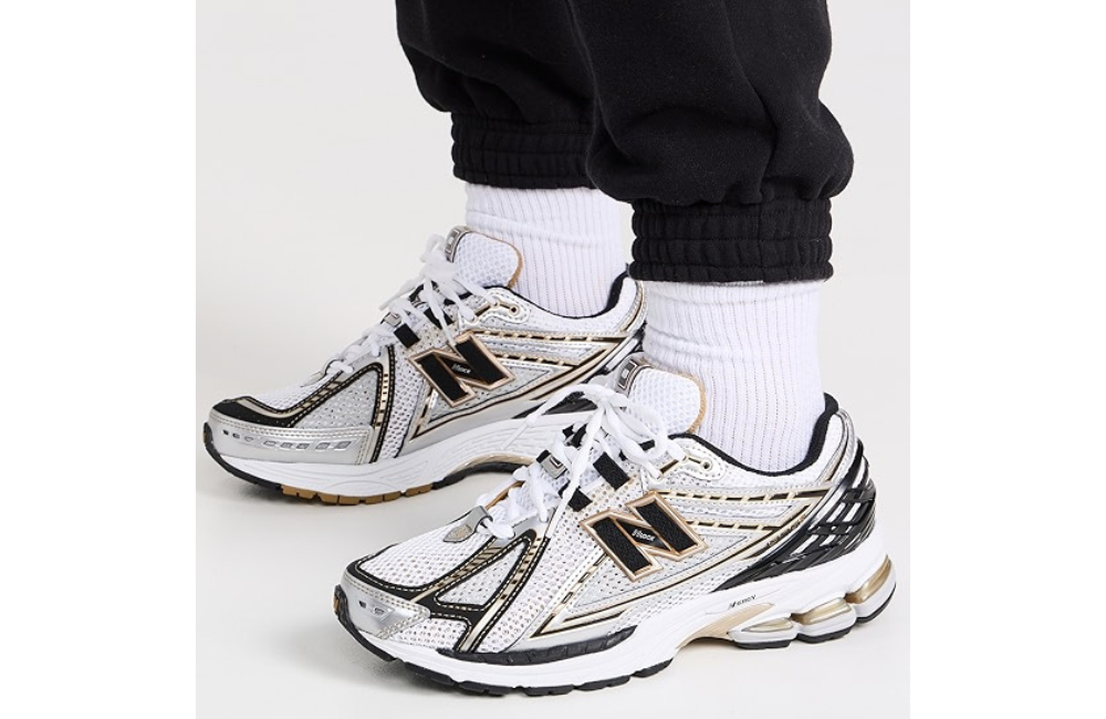A side view of a person wearing New Balance 1906R sneakers in white with gold and black accents. The shoes have a sporty, modern design with detailed overlays. They are paired with plain white socks and black jogger pants.