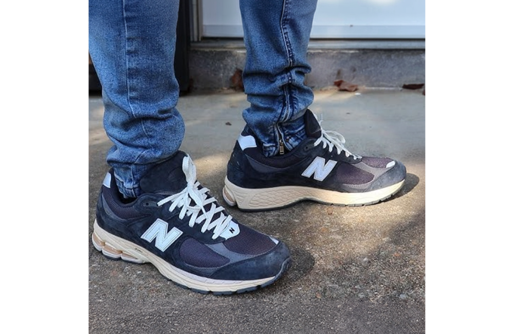 A close-up of a person wearing New Balance 2002R sneakers in a black and dark grey colorway. The shoes feature a rugged sole and a combination of mesh and suede materials. The sneakers are styled with light blue jeans.