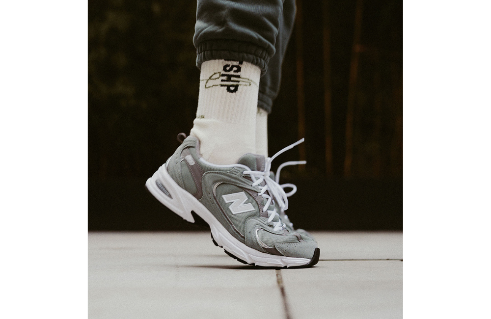 A close-up view of a person wearing New Balance 530 sneakers in grey (Juniper Shadow) with white accents. The shoes have a classic, retro-inspired design and are paired with white socks that have bold black text, as well as grey jogger pants.
